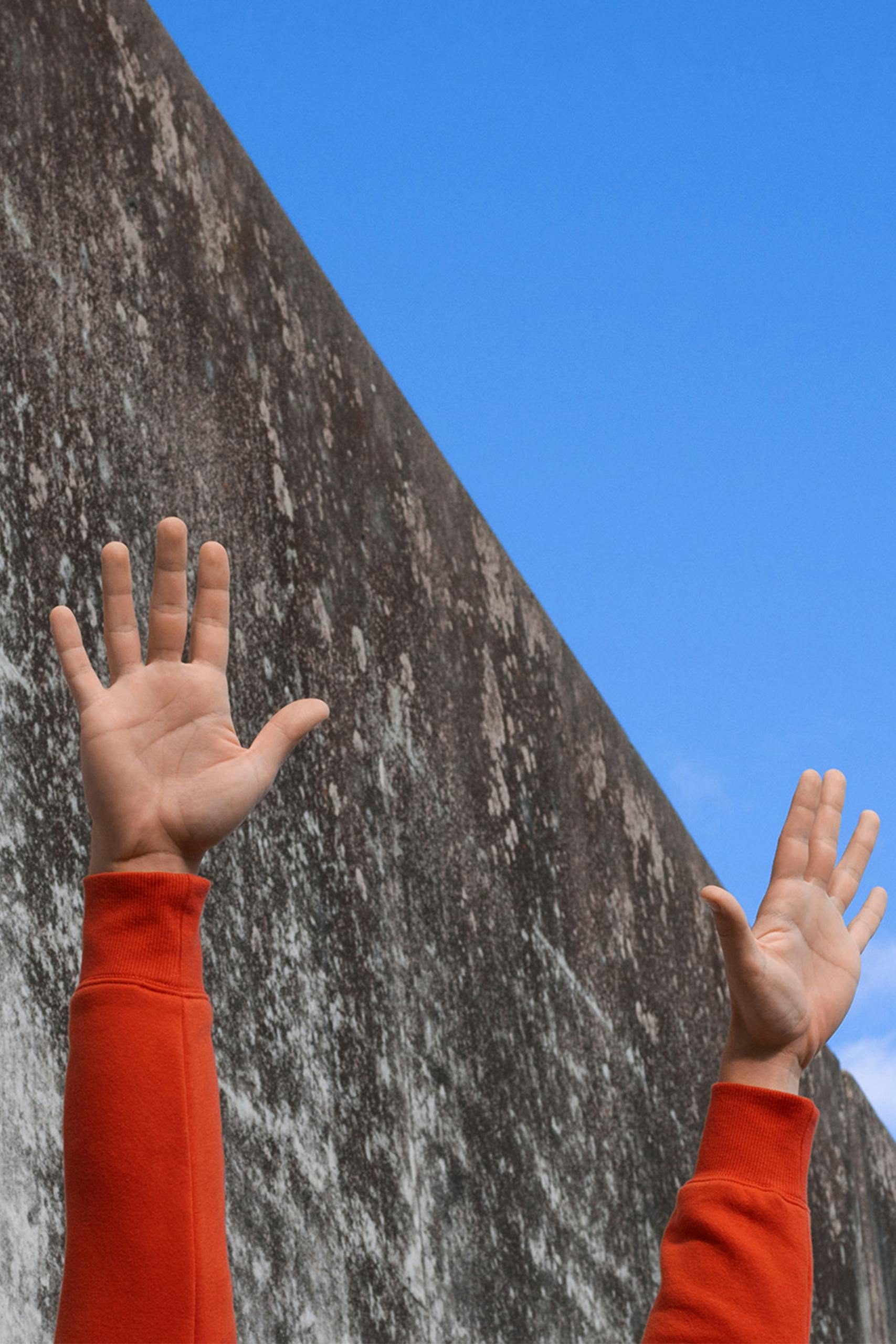 Hands in orange sleeves reaching towards blue sky, against a concrete wall.